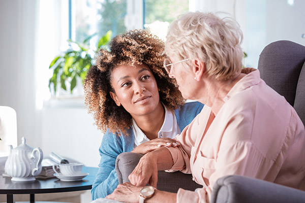 Female home caregiver talking with senior woman, sitting in living room and listening to her carefully.